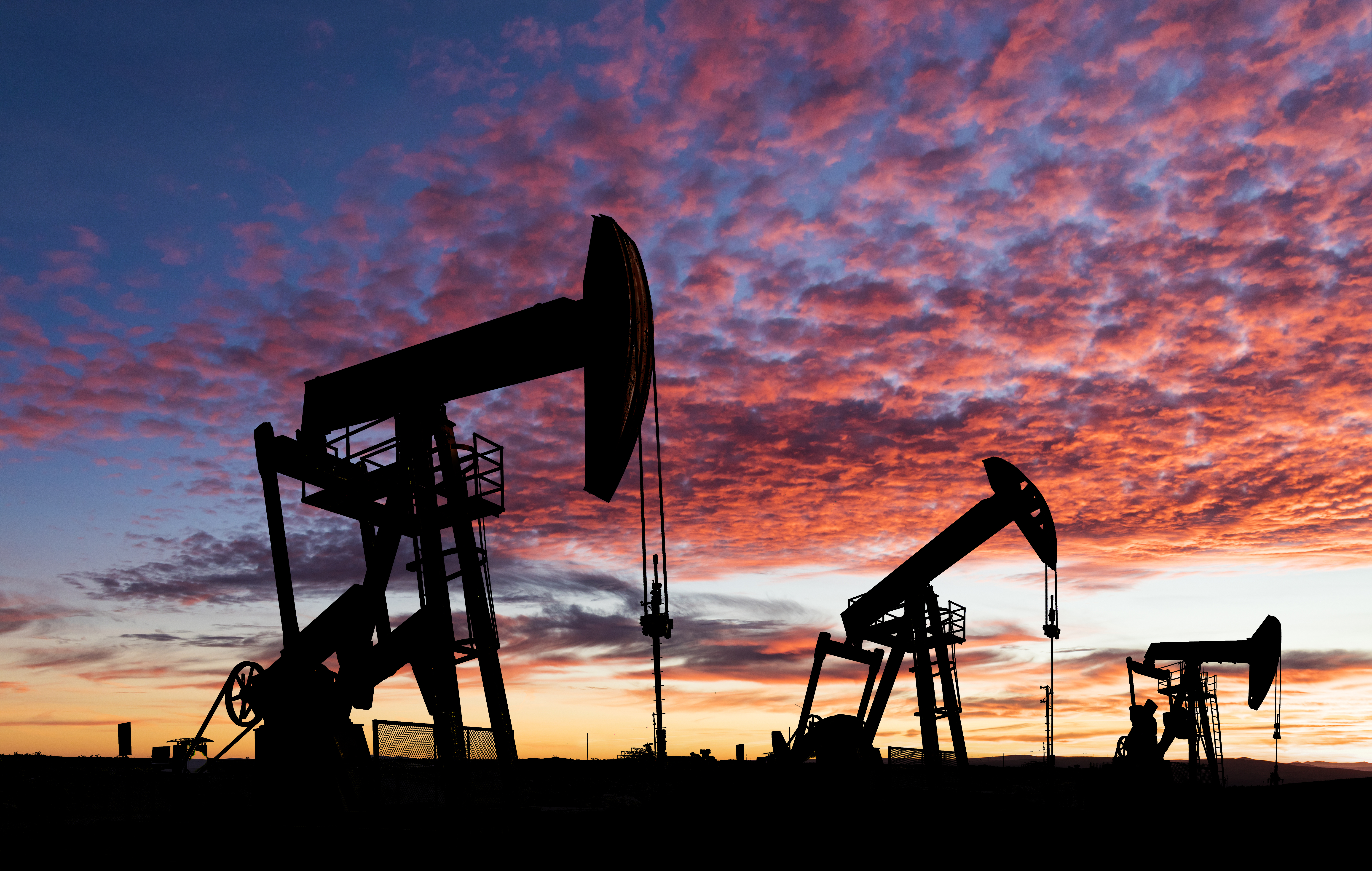 3 Silhouettes of oil pump jacks stand against a vibrant sunset sky with scattered clouds.