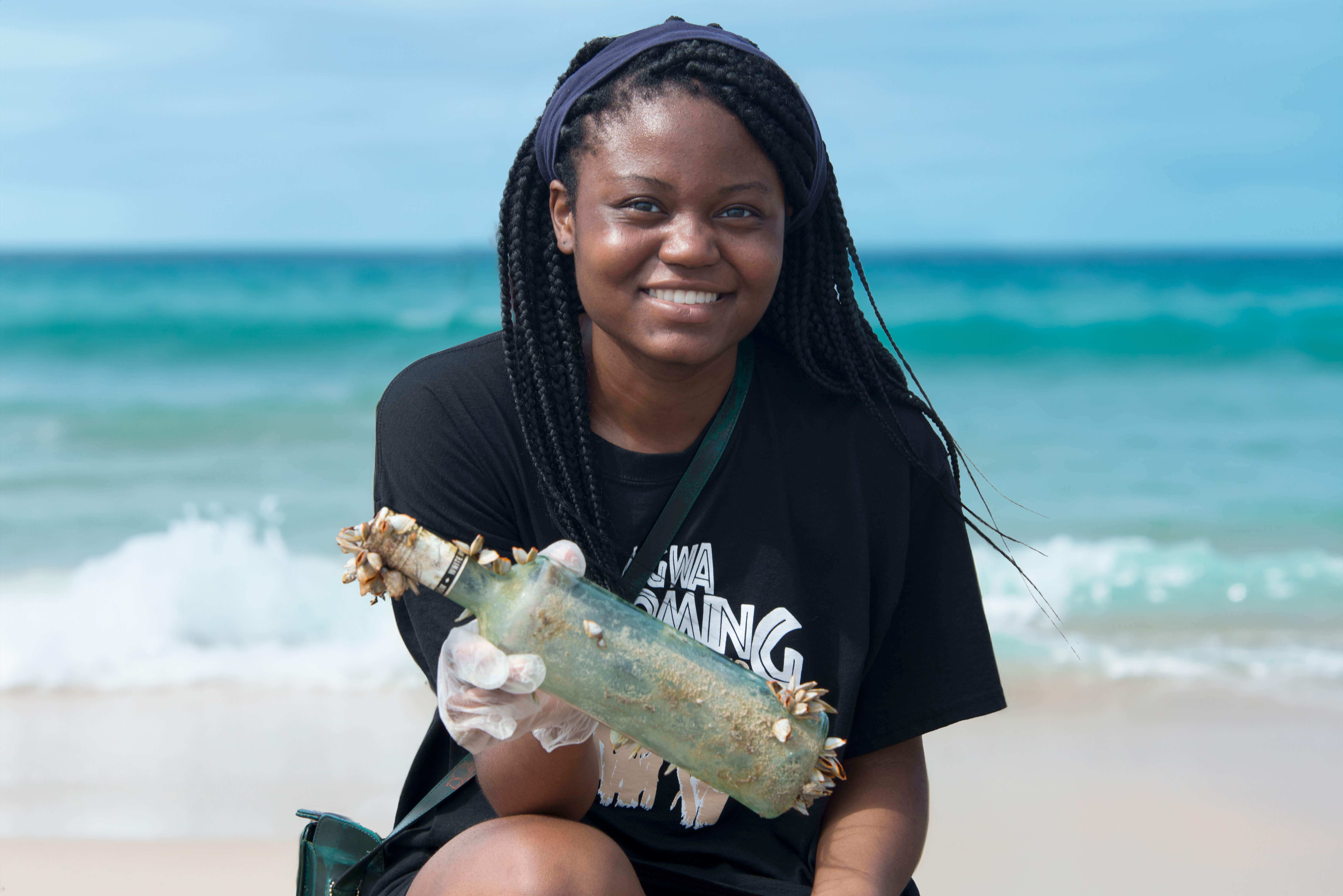 An African American girl holds up a bottle with mollusks on it that she found on the beach with the ocean in the background.