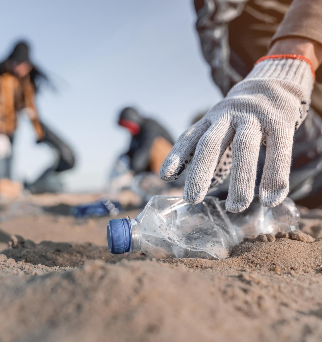 Close up of a hand picking up a plastic bottle on the beach with other volunteers in the background.