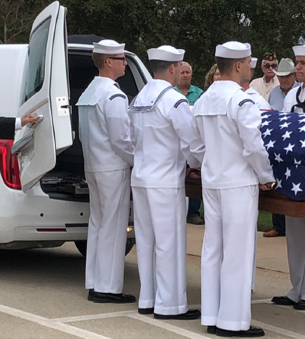 A casket covered in an American flag being carried out of a white hearse by 6 American Navy sailors.