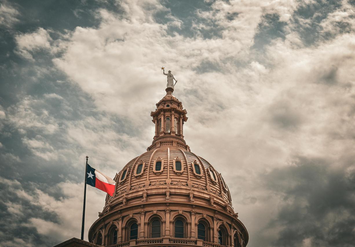 Looking up at the dome of the Texas Capitol building with Texas flag and scattered clouds in sky.
