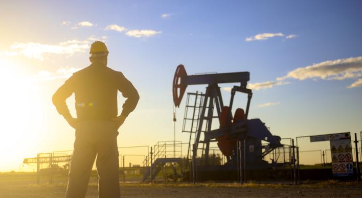 Silhouette of male worker with hardhat overlooking a pumpjack with bright sun in the background